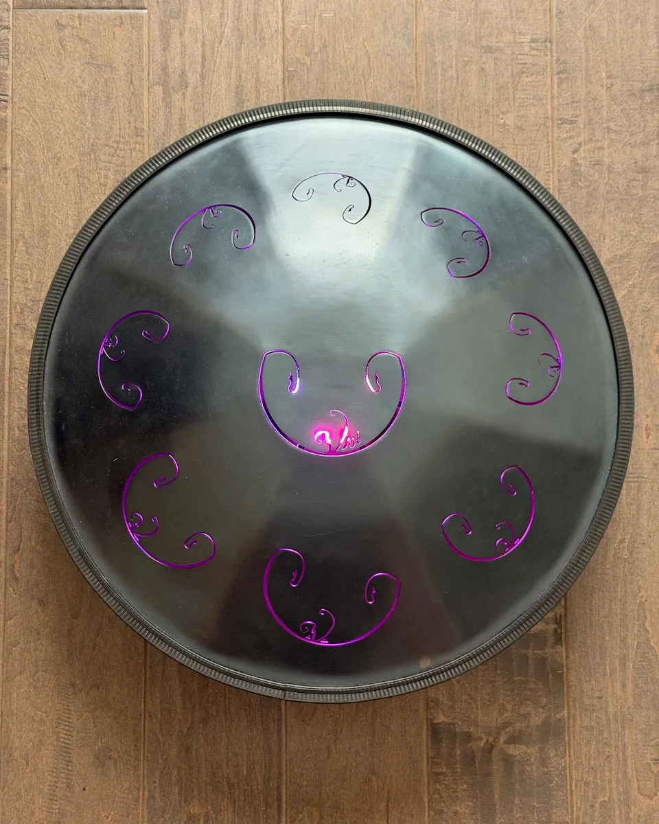 Top-down view of a black RAV Vast tongue drum on a wood floor, its tongue cutouts glowing purple and magenta from the Aura Light beneath