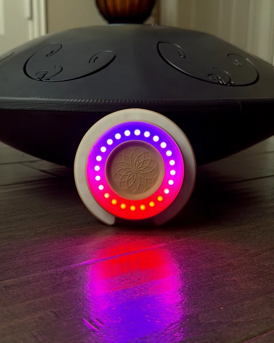 Aura Light standing on a wood floor in front of a black RAV Vast tongue drum, its LED ring glowing purple, pink, red, and amber and casting a colorful reflection